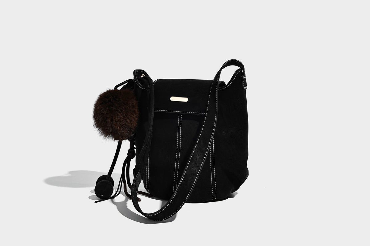 Small matte textured bucket crossbody bag with flap closure, gold-tone bar hardware, contrast stitching, and pompom tassel