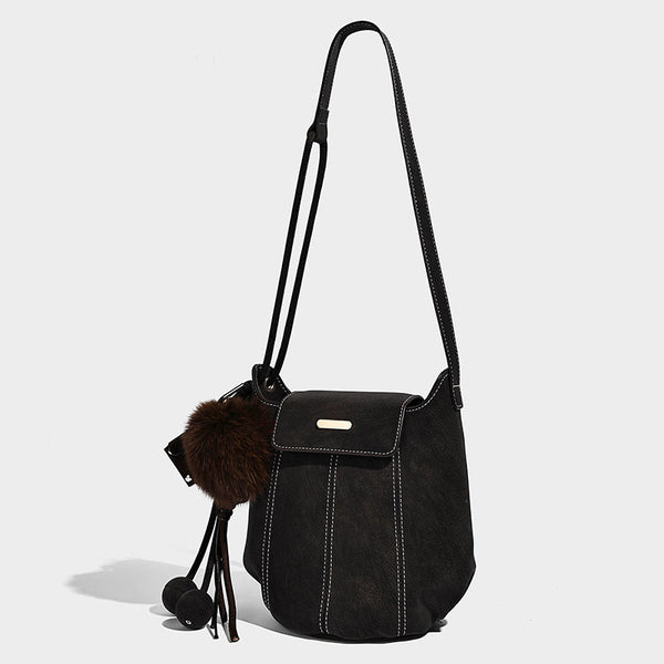 Small matte textured bucket crossbody bag with flap closure, gold-tone bar hardware, contrast stitching, and pompom tassel