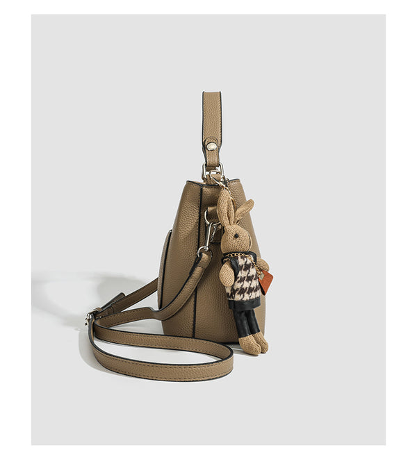 Structured taupe pebbled-texture bucket bag with top handle, silver ring hardware, tab-and-button closure, and knit bunny