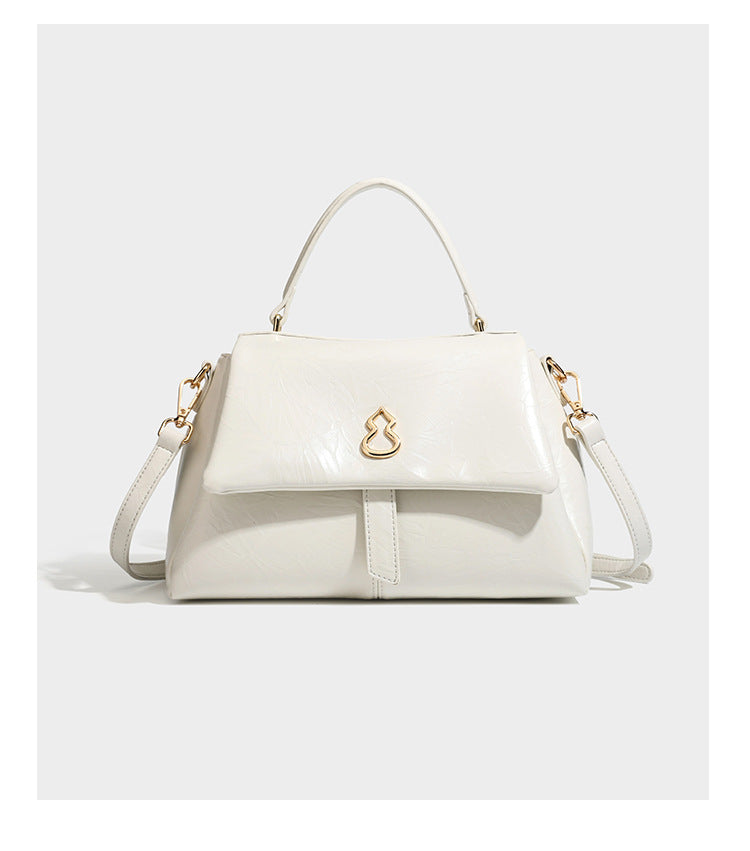 Structured black satchel bag with glossy crinkled leather-look finish, gold-tone emblem flap closure, slim top handle, and