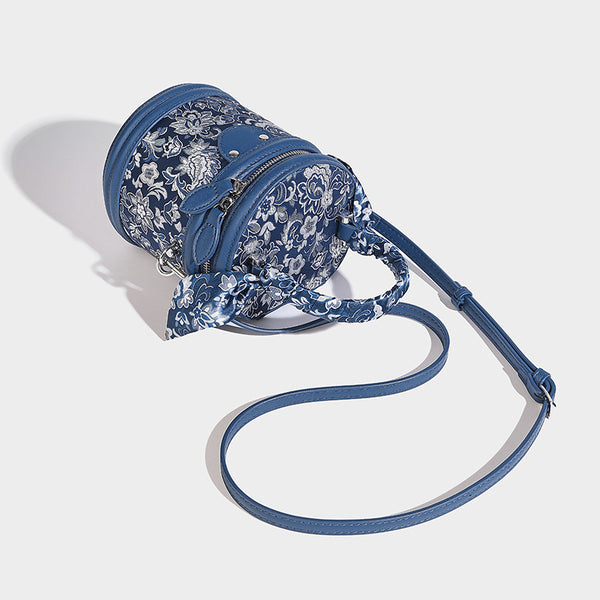 Small cylindrical barrel bag in blue-and-white floral jacquard with leather-look trim, scarf-wrapped top handle, silver-tone