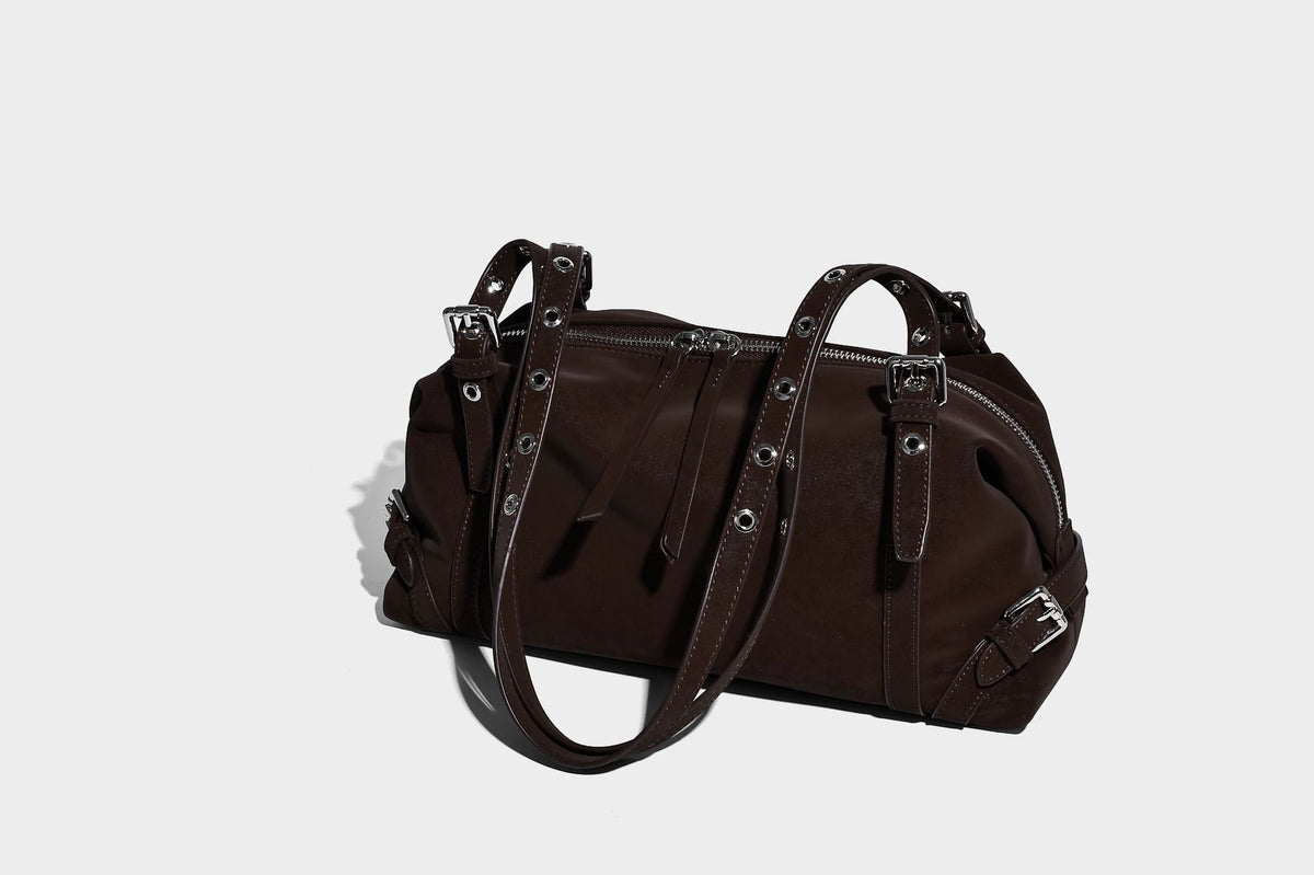 Weiche, zylinderförmige Schultertasche in Dunkelbraun mit silberfarbenen Riemen mit Ösen, Reißverschluss oben, seitlichen Schnallenbeschlägen und Quaste