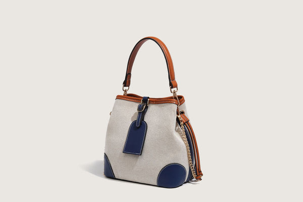 Cream woven-texture canvas bucket bag with navy accent panels, tan leather-look trim, gold-tone chain strap, and tassel