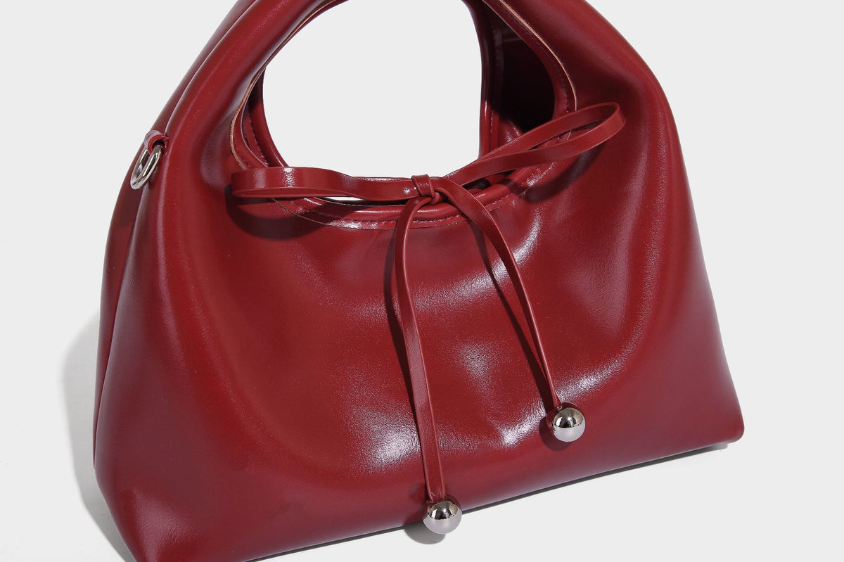 Kleine kuppelförmige Henkeltasche in glänzender Lederoptik mit Kordelzugverschluss, weißem Spitzen-Schleifenanhänger und silberfarbenen Metallbeschlägen
