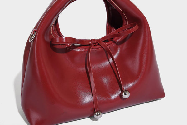 Kleine kuppelförmige Henkeltasche in glänzender Lederoptik mit Kordelzugverschluss, weißem Spitzen-Schleifenanhänger und silberfarbenen Metallbeschlägen