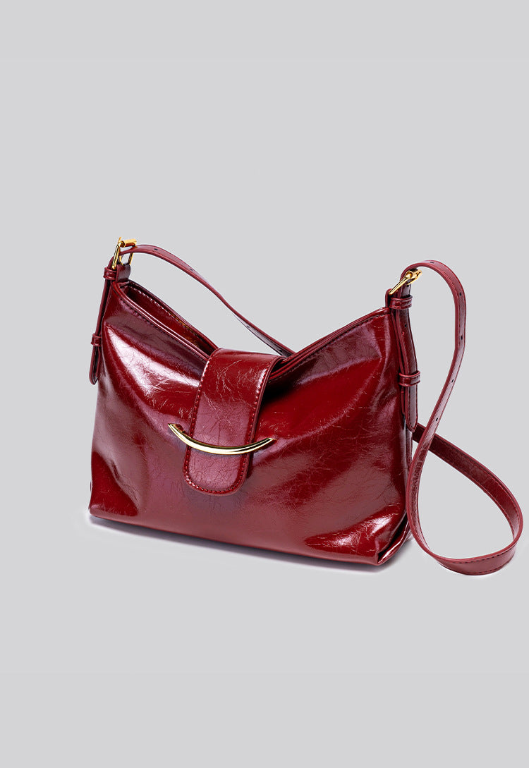 Glossy burgundy leather-look shoulder bag with curved gold-tone bar front closure, crinkled oil-wax texture, and adjustable