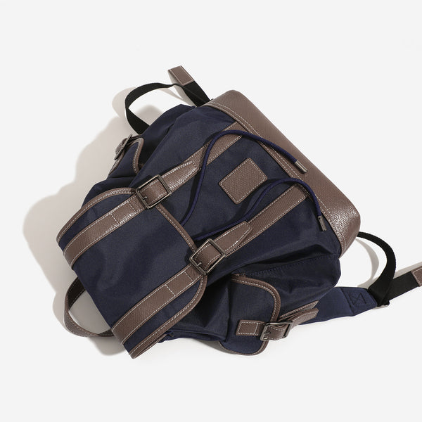 Deep navy canvas backpack with brown textured leather-look trim, drawstring flap closure, buckle-strap front detail, and