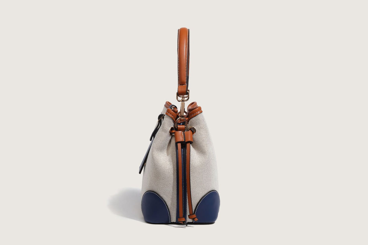 Cream woven-texture canvas bucket bag with navy accent panels, tan leather-look trim, gold-tone chain strap, and tassel