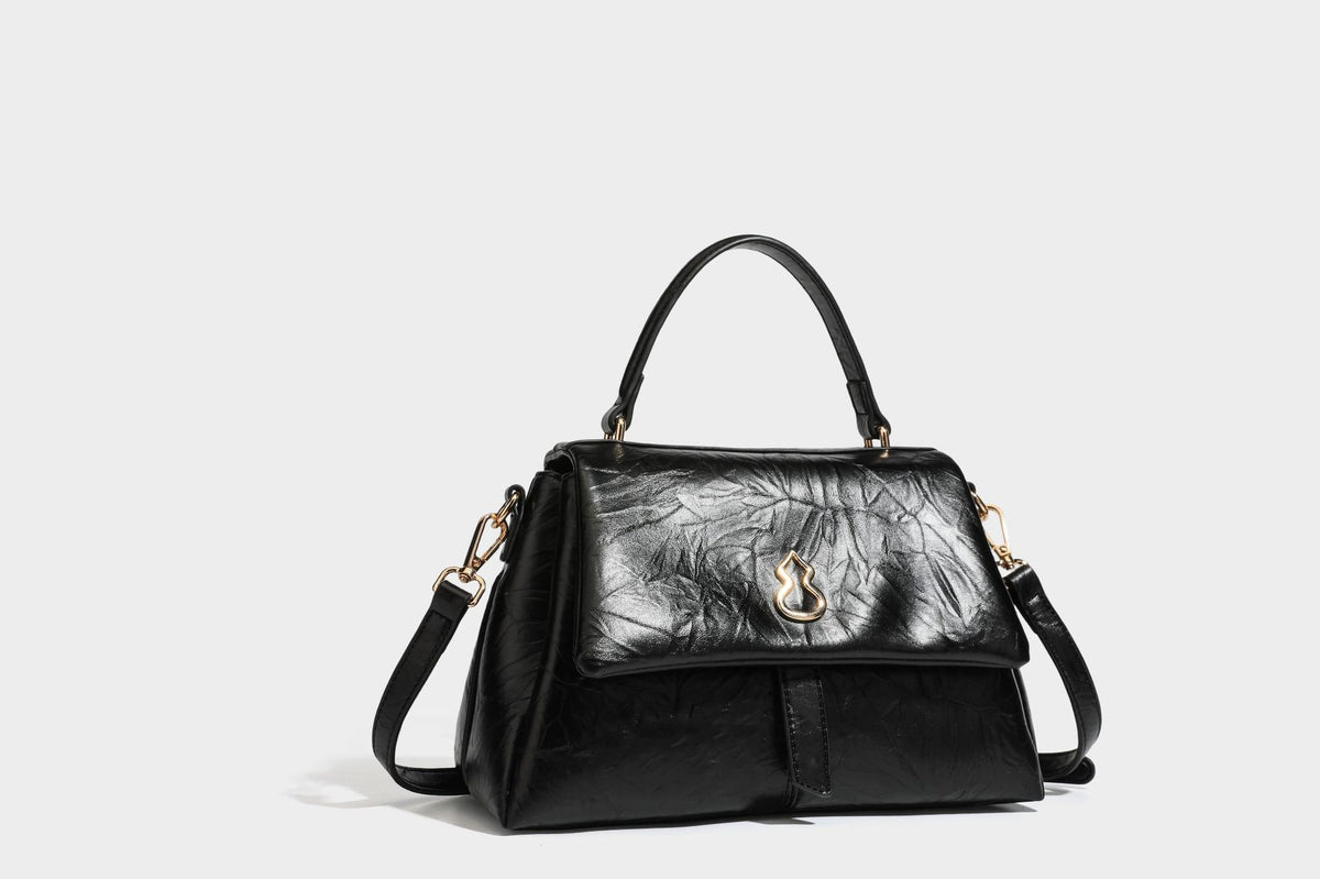 Structured black satchel bag with glossy crinkled leather-look finish, gold-tone emblem flap closure, slim top handle, and