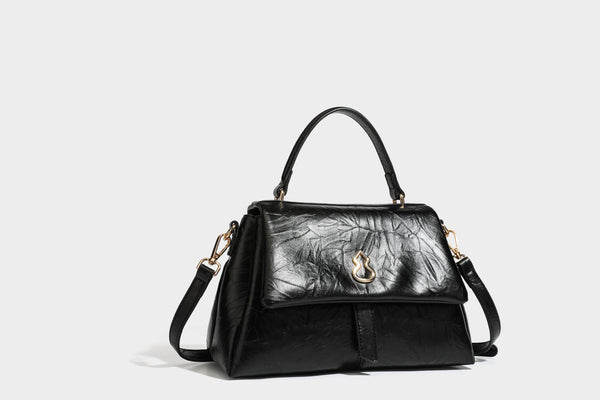 Structured black satchel bag with glossy crinkled leather-look finish, gold-tone emblem flap closure, slim top handle, and