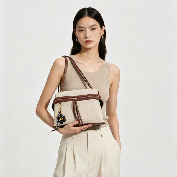Cream canvas tote bag with brown leather-look trim, dual exterior zip pockets, long shoulder strap, and black flower charm