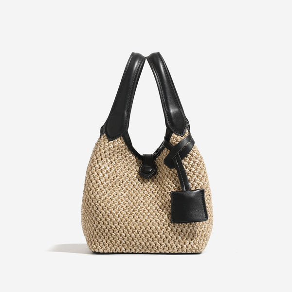 Woven beige straw bucket bag with black leather-look trim, dual top handles, detachable shoulder strap, and front snap