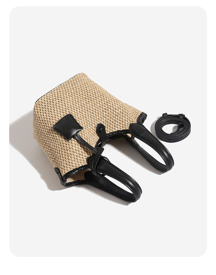 Woven beige straw bucket bag with black leather-look trim, dual top handles, detachable shoulder strap, and front snap