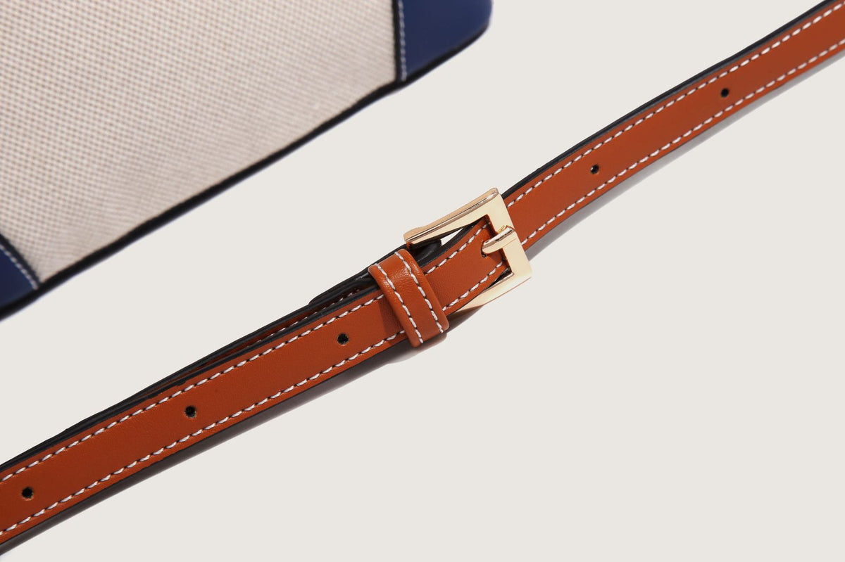 Cream woven-texture canvas bucket bag with navy accent panels, tan leather-look trim, gold-tone chain strap, and tassel