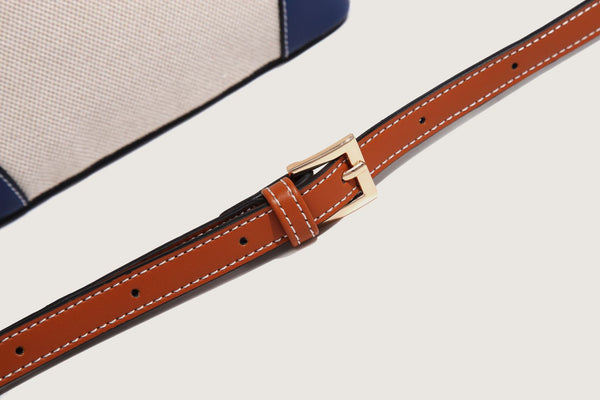 Cream woven-texture canvas bucket bag with navy accent panels, tan leather-look trim, gold-tone chain strap, and tassel
