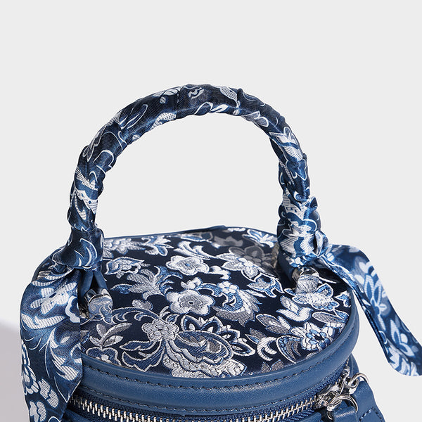 Small cylindrical barrel bag in blue-and-white floral jacquard with leather-look trim, scarf-wrapped top handle, silver-tone