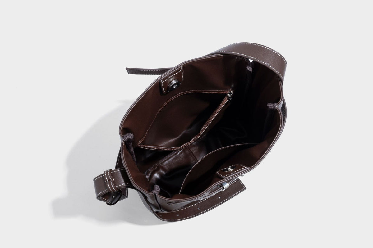 Structured dark brown leather-look bucket bag with white contrast stitching, front wraparound belt buckle strap, and long