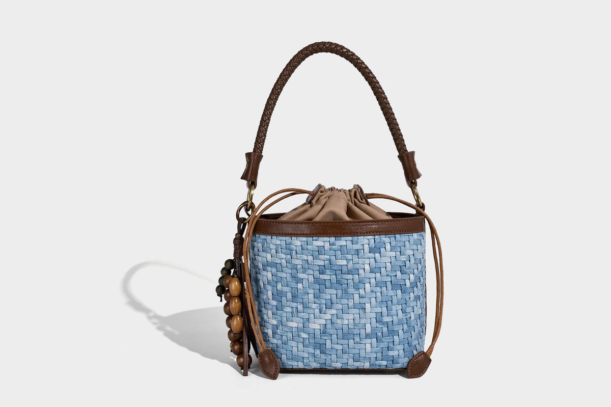 Small light blue woven bucket bag with brown leather-look trim, braided top handle, brass-tone ring hardware, drawstring