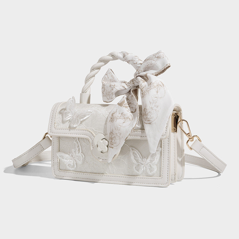 Small structured white flap bag with butterfly embroidery, flower-shaped turn-lock clasp, braided top handle with scarf bow