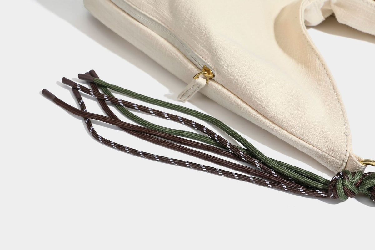 Soft slouchy cream canvas shoulder tote with braided brown and green cord handle, gold-tone ring hardware, and tassel trim