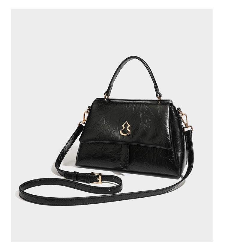 Structured black satchel bag with glossy crinkled leather-look finish, gold-tone emblem flap closure, slim top handle, and