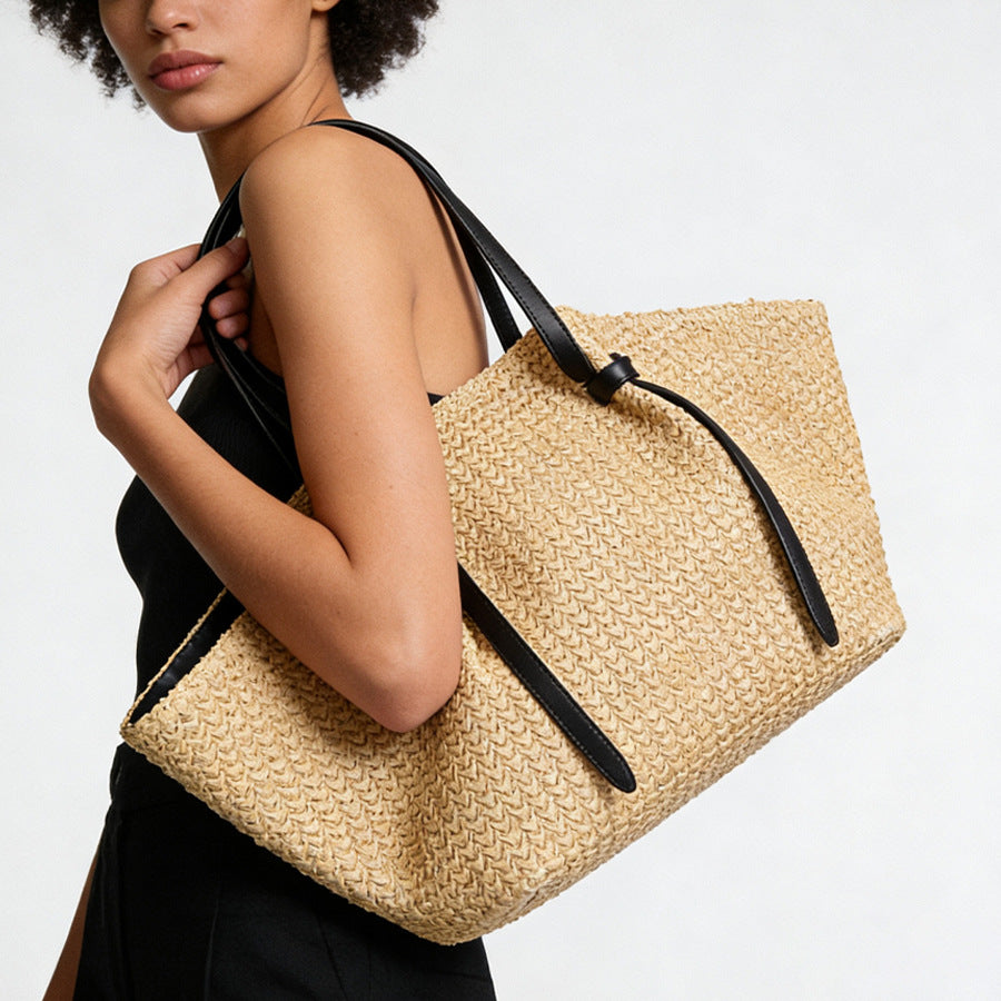 Large woven straw tote bag with black leather-look shoulder straps and knotted strap detail, natural tan trapezoid body