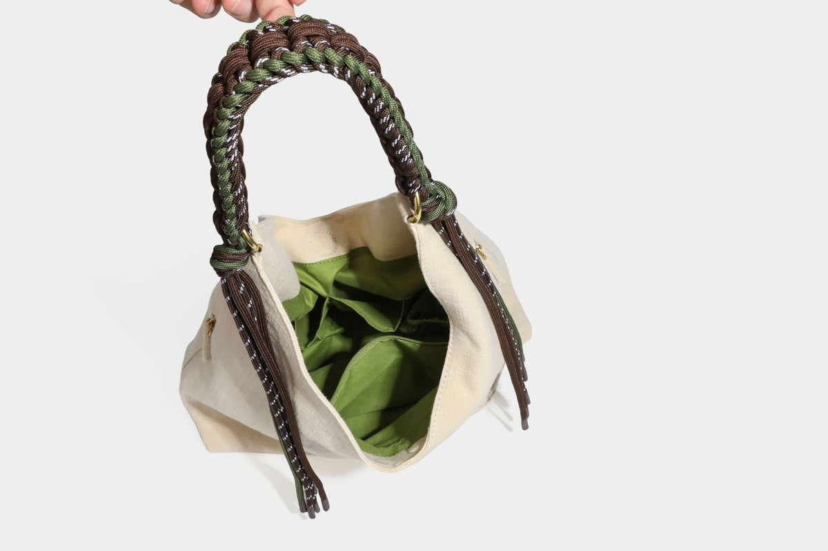 Soft slouchy cream canvas shoulder tote with braided brown and green cord handle, gold-tone ring hardware, and tassel trim