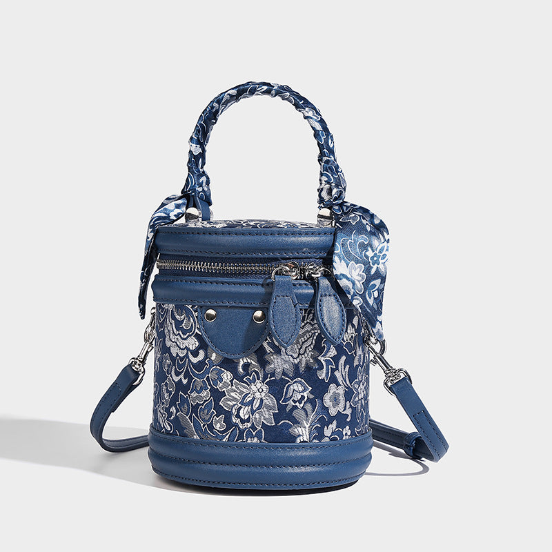 Small cylindrical barrel bag in blue-and-white floral jacquard with leather-look trim, scarf-wrapped top handle, silver-tone