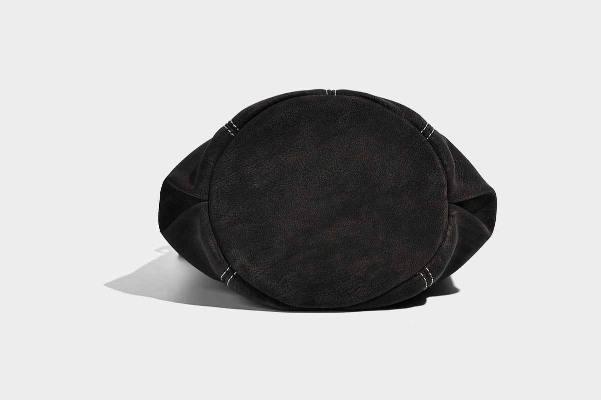 Small matte textured bucket crossbody bag with flap closure, gold-tone bar hardware, contrast stitching, and pompom tassel