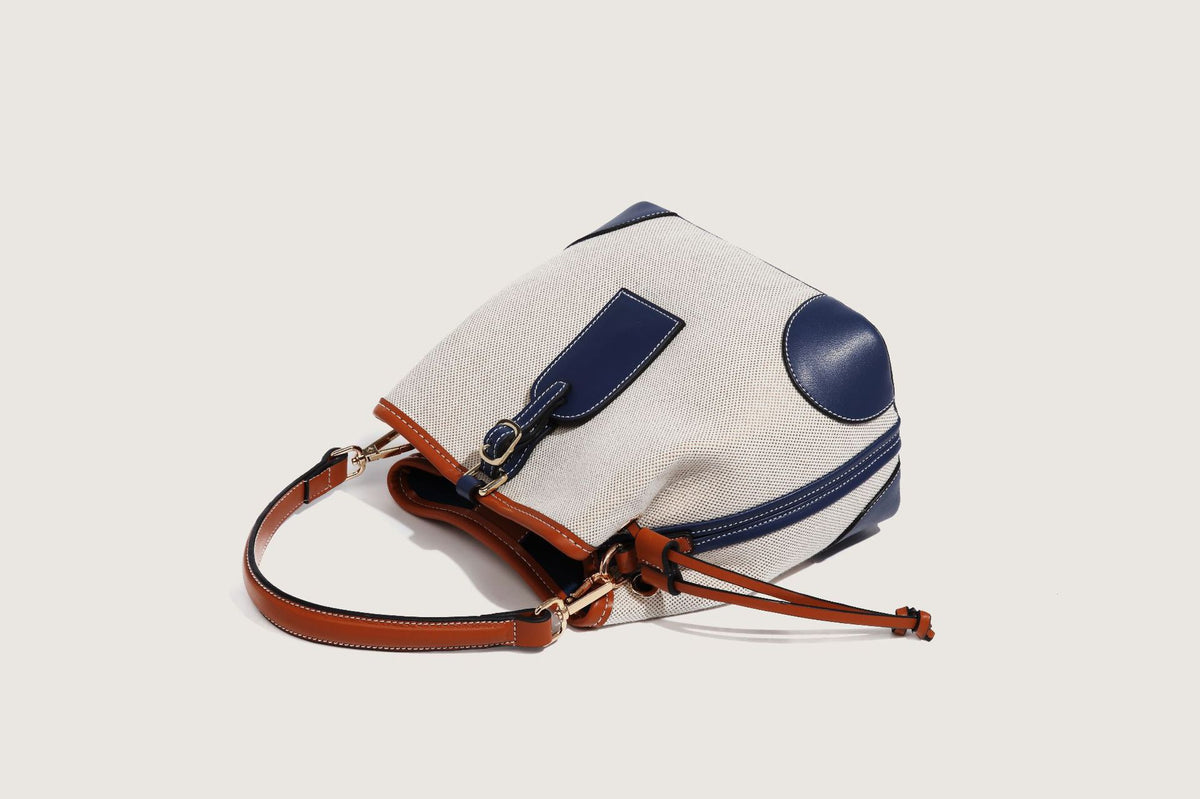 Cream woven-texture canvas bucket bag with navy accent panels, tan leather-look trim, gold-tone chain strap, and tassel