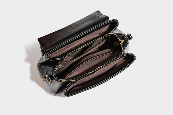 Structured black satchel bag with glossy crinkled leather-look finish, gold-tone emblem flap closure, slim top handle, and