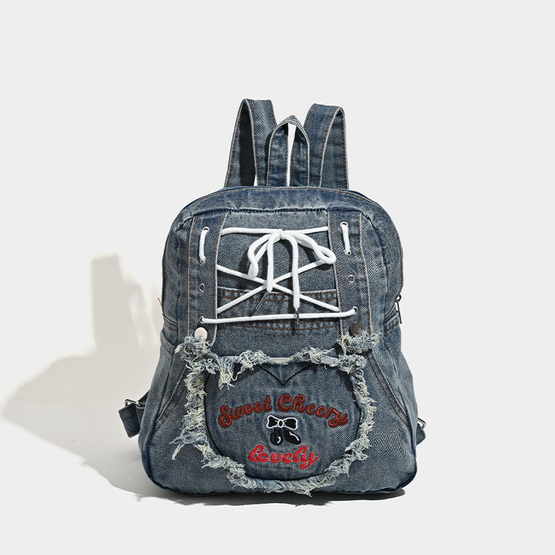 Washed denim blue backpack with white lace-up cord front panel, frayed heart-shaped embroidered exterior pocket, zip top