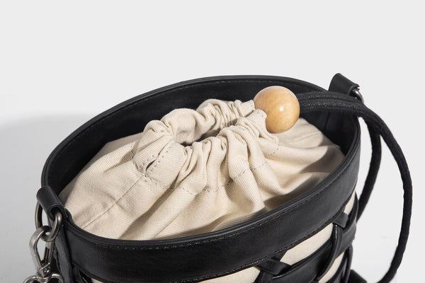 Small cylindrical woven lattice bucket bag with leather-look black trim, beige canvas body, wooden bead drawstring closure