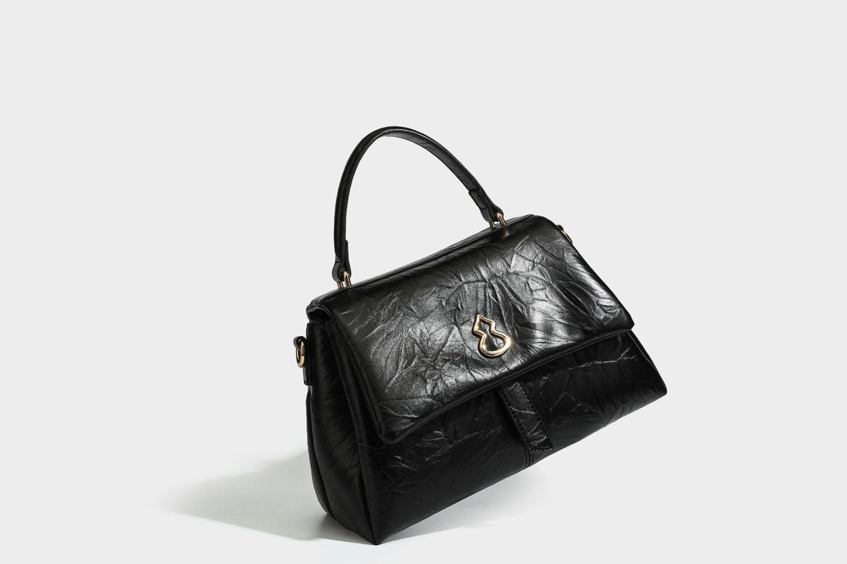 Structured black satchel bag with glossy crinkled leather-look finish, gold-tone emblem flap closure, slim top handle, and