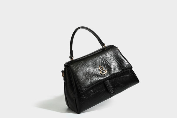 Structured black satchel bag with glossy crinkled leather-look finish, gold-tone emblem flap closure, slim top handle, and