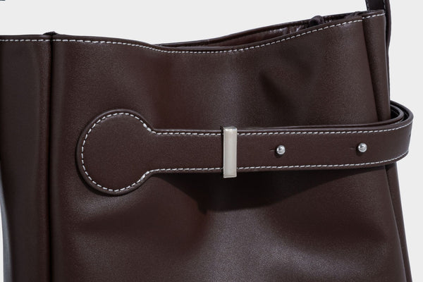 Structured dark brown leather-look bucket bag with white contrast stitching, front wraparound belt buckle strap, and long
