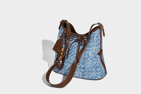 Light blue woven bucket bag with brown leather-look trim, gold-tone hardware, adjustable shoulder strap, and decorative bead