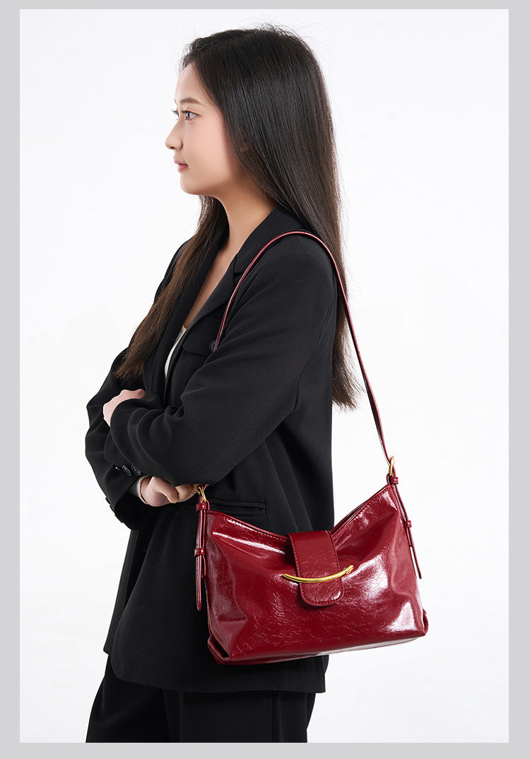 Glossy burgundy leather-look shoulder bag with curved gold-tone bar front closure, crinkled oil-wax texture, and adjustable