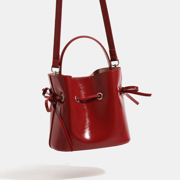 Compact glossy leather-look bucket bag with drawstring bow-tie closure, rounded top handle, and silver-tone grommet hardware