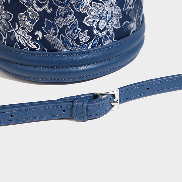Small cylindrical barrel bag in blue-and-white floral jacquard with leather-look trim, scarf-wrapped top handle, silver-tone
