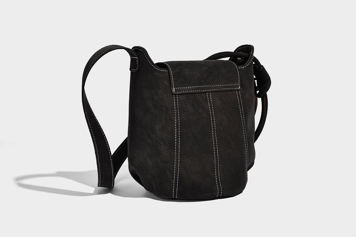 Small matte textured bucket crossbody bag with flap closure, gold-tone bar hardware, contrast stitching, and pompom tassel