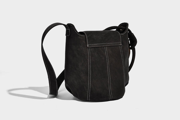 Small matte textured bucket crossbody bag with flap closure, gold-tone bar hardware, contrast stitching, and pompom tassel