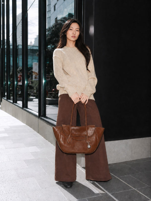 Large winged leather-look tote bag with single arched handle, stitched flap-front panel, and silver ball charm in cognac