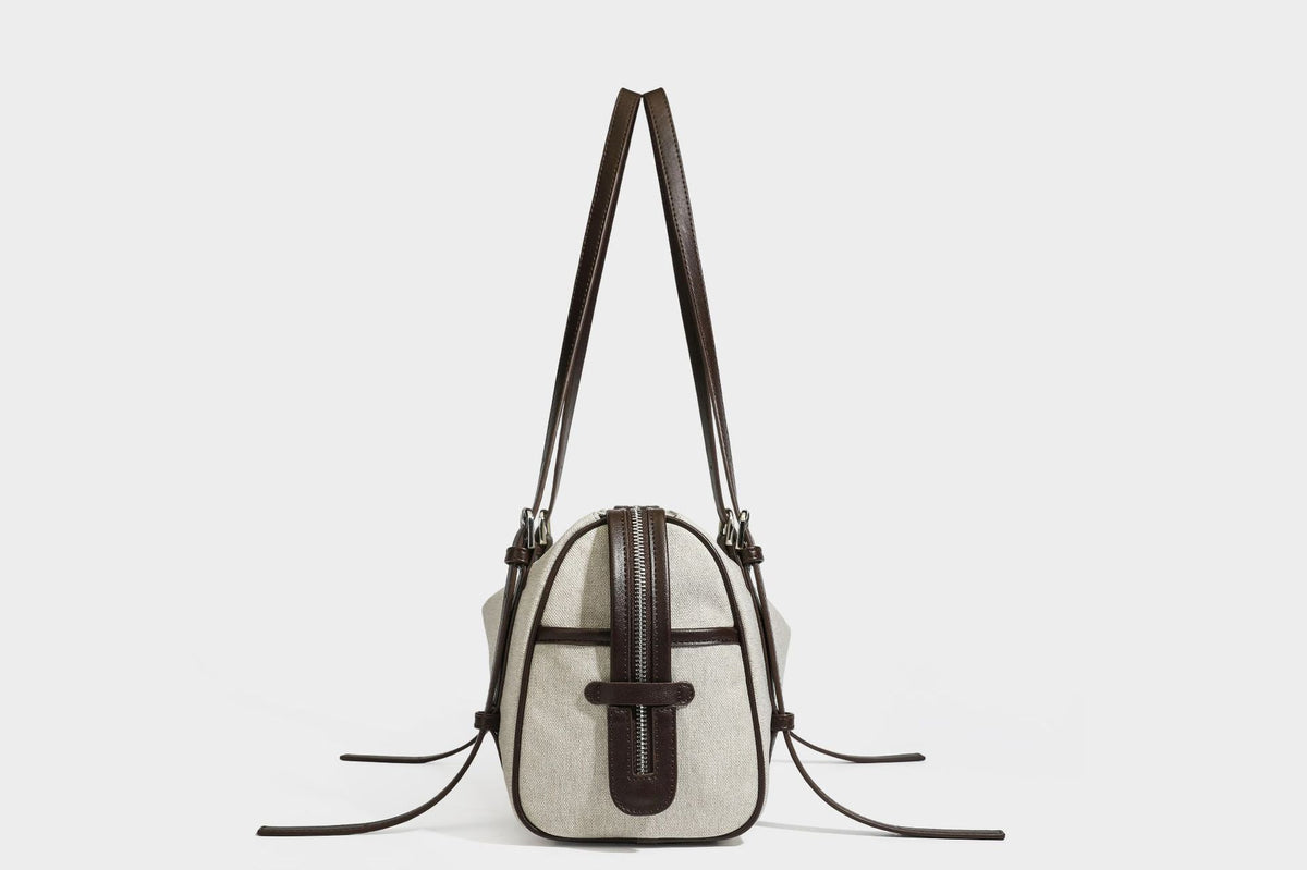 Beige canvas barrel bag with dark brown leather-look trim, silver-tone hardware, top zip closure, and adjustable shoulder