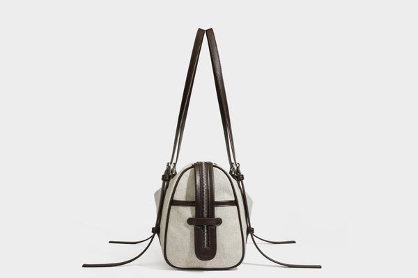 Beige canvas barrel bag with dark brown leather-look trim, silver-tone hardware, top zip closure, and adjustable shoulder