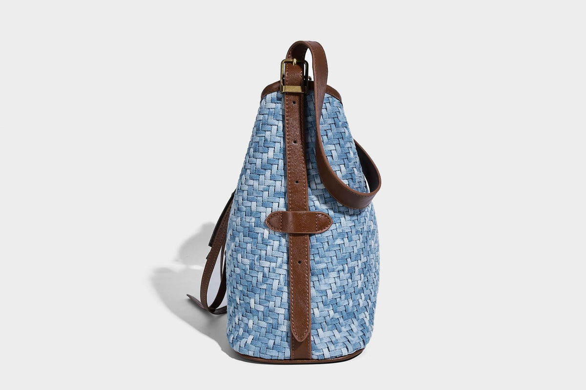 Light blue woven bucket bag with brown leather-look trim, gold-tone hardware, adjustable shoulder strap, and decorative bead