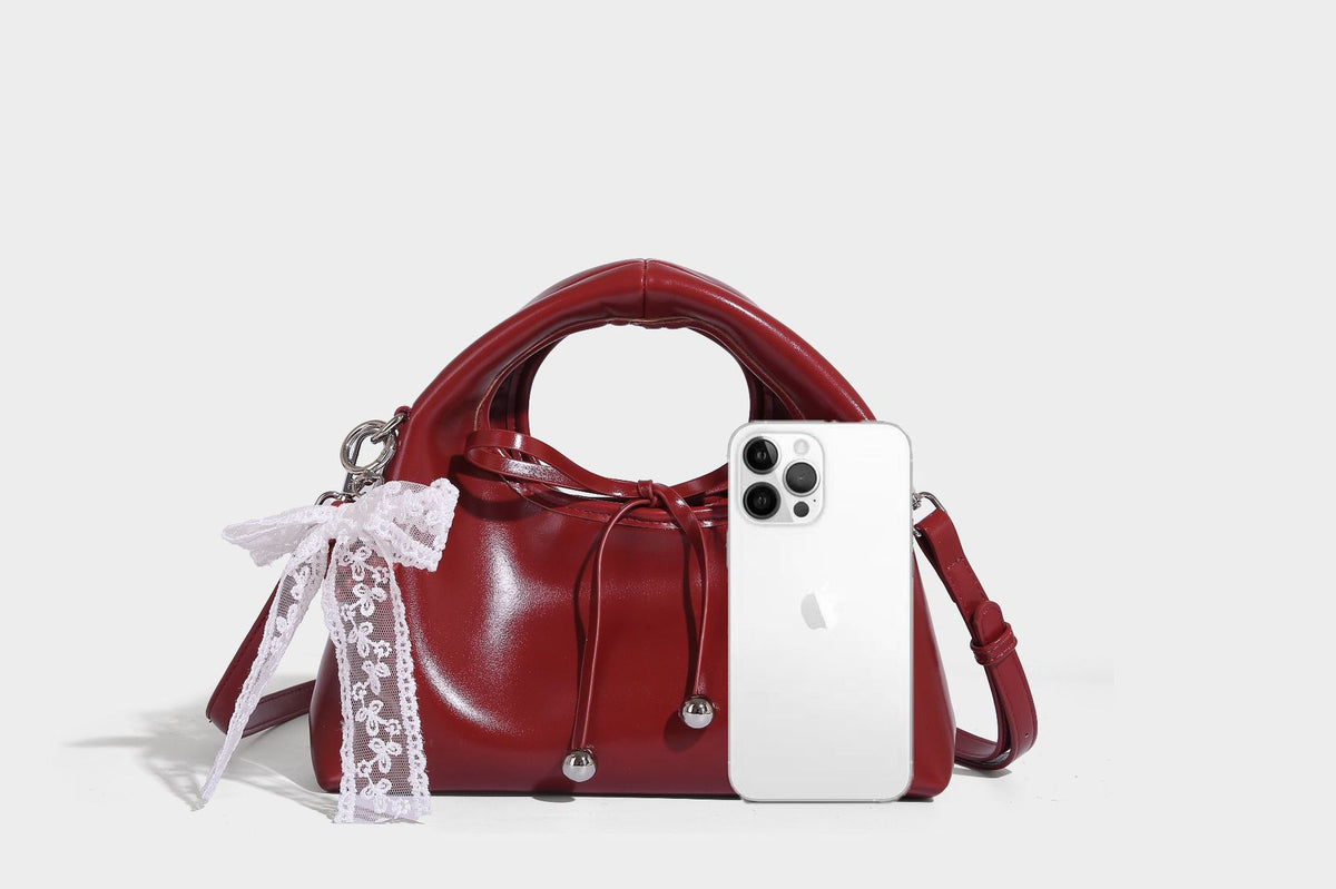 Kleine kuppelförmige Henkeltasche in glänzender Lederoptik mit Kordelzugverschluss, weißem Spitzen-Schleifenanhänger und silberfarbenen Metallbeschlägen