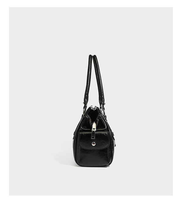 Glänzende, geknautschte Barrel-Satchel mit zwei oberen Tragegriffen, vorderem Klappen-Riemen-Panel, Ring- und Nietenbeschlägen sowie einem abnehmbaren