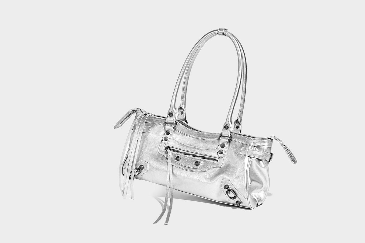 Längliche Schultertasche in Zylinderform mit glänzender Lederoptik, silberfarbenen Nietenbeschlägen, seitlichen Quasten und mehreren außenliegenden Reißverschlüssen