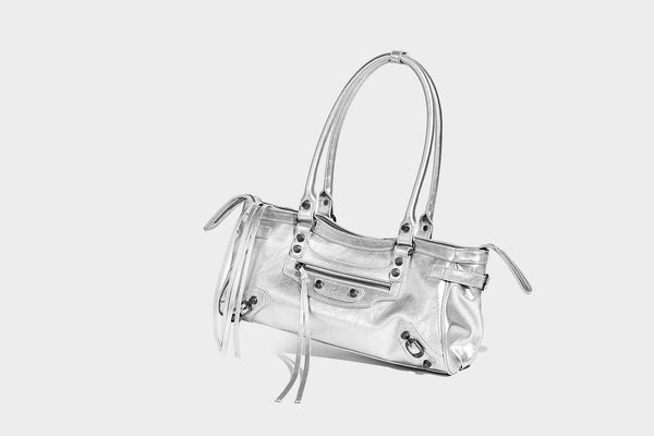 Längliche Schultertasche in Zylinderform mit glänzender Lederoptik, silberfarbenen Nietenbeschlägen, seitlichen Quasten und mehreren außenliegenden Reißverschlüssen